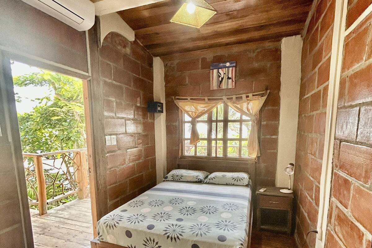 Habitació de fusta amb un llit, una finestra amb cortines, una tauleta de nit i un aparell d'aire condicionat.