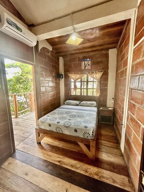 Una habitación de madera con una cama, una ventana con cortinas, una mesita de noche y un aire acondicionado.