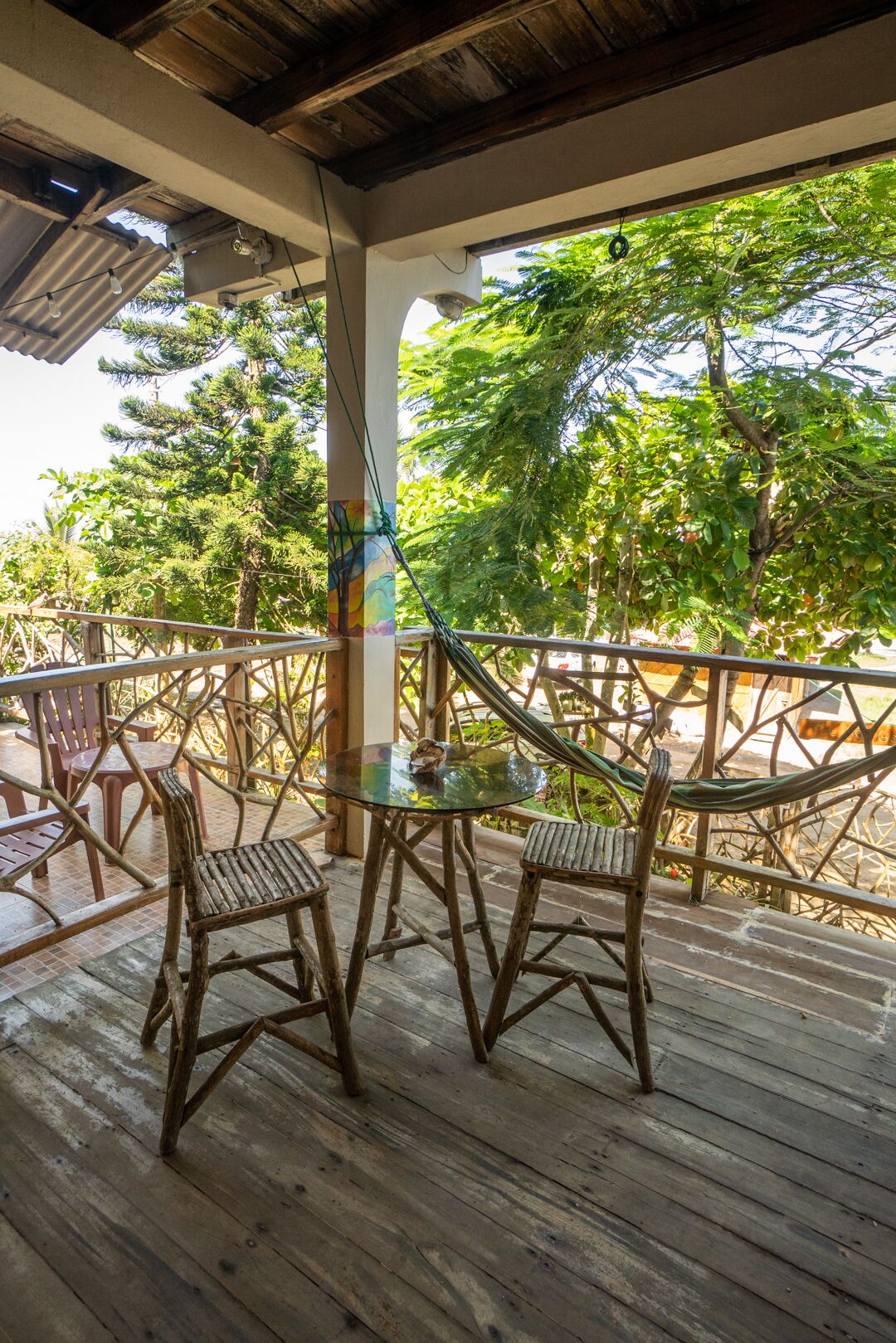 Una terraza de madera con mobiliario exterior, que incluye sillas y una mesa, con vistas a un jardín exuberante con árboles y un cielo despejado.
