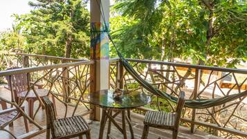 Una terraza de madera con mobiliario exterior, que incluye sillas y una mesa, con vistas a un jardín exuberante con árboles y un cielo despejado.