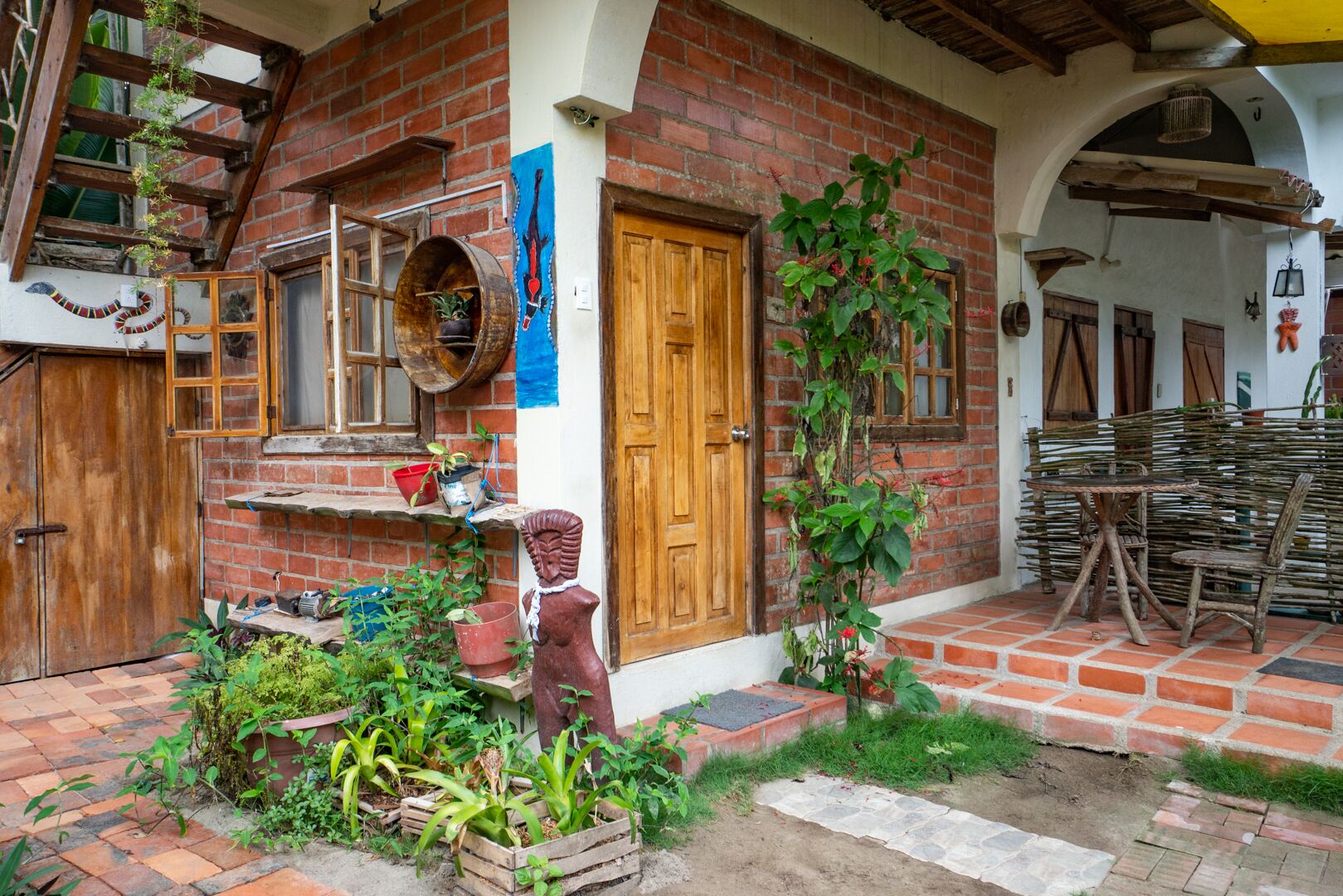 Un edificio rústico de ladrillo con una puerta de madera, un pequeño jardín con plantas y una escultura de madera, y un juego de mesa y silla de mimbre.