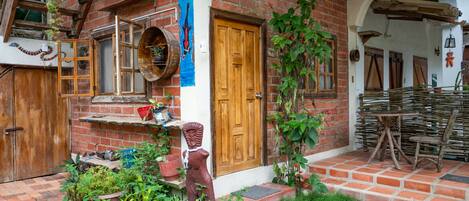Un edificio rústico de ladrillo con una puerta de madera, un pequeño jardín con plantas y una escultura de madera, y un juego de mesa y silla de mimbre.