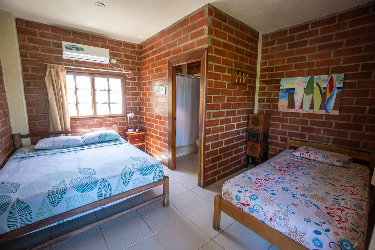Una habitació amb dos llits, una finestra amb cortines, una paret de maó i un quadre de taules de surf.