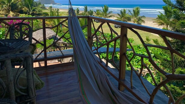 Un hamac sur une terrasse en bois donnant sur une plage bordée de palmiers, sous un ciel dégagé.