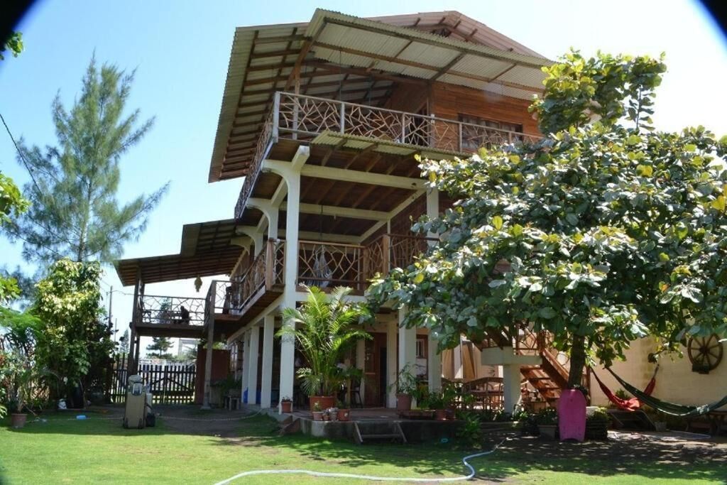 A two-story house with a balcony, surrounded by greenery and a hammock.
