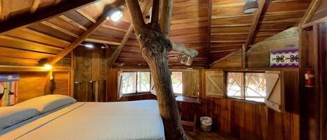 A rustic wooden room with a bed, a large tree trunk, and a window with shutters.