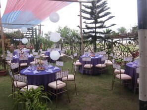 Un espacio al aire libre para eventos con mesas redondas cubiertas con manteles azules, sillas blancas y farolillos de papel blancos.