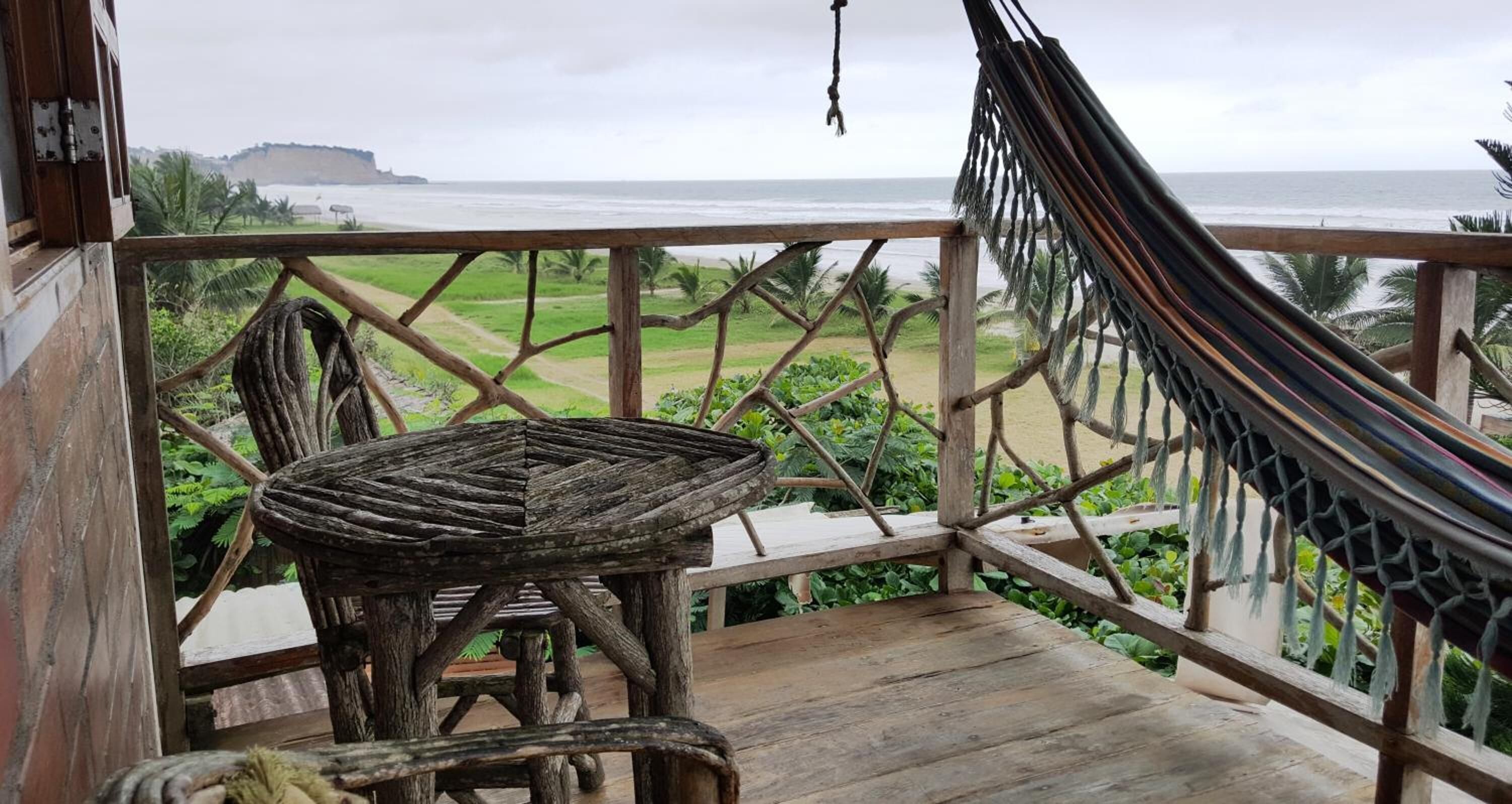 A wooden deck with a hammock and a rustic table, overlooking a beach and a distant cliff.