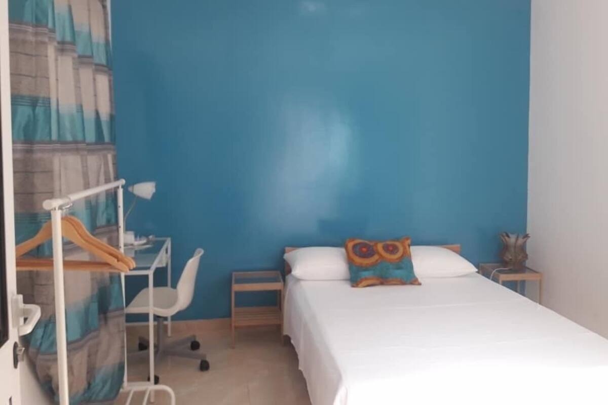 A bedroom with a bed, a desk, a chair, a hanging rack, and a nightstand.