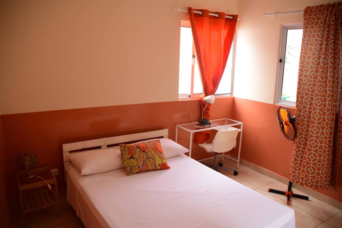A bedroom with a bed, a desk, a chair, a standing fan, and a window with curtains.