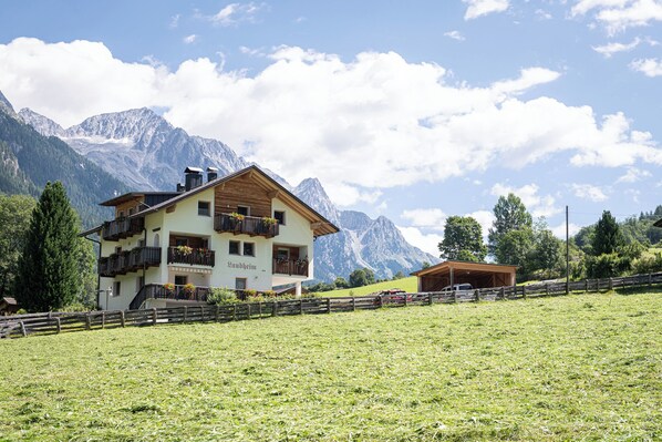Ein mehrstöckiges Gebäude mit Balkonen, umgeben von einem Holzzaun, vor einer Bergkulisse und blauem Himmel.