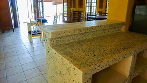 A kitchen area with a granite countertop, tiled floor, and a dining area with chairs and a table. - Casa Ruth (San Juan del Sur)