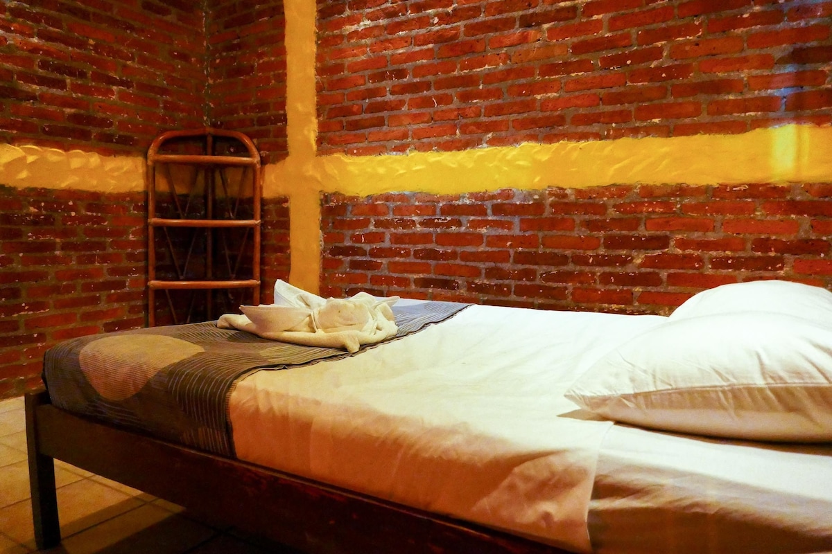 Uma cama de solteiro com estrutura de madeira, uma parede de tijolos e uma escada de metal encostada na parede.