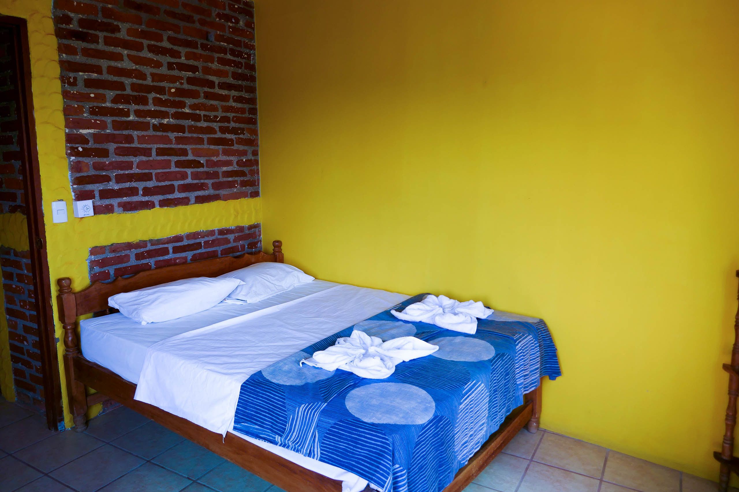 Un lit simple avec du linge de lit blanc et une couvre-lit bleue à motifs, placé devant un mur jaune dont la partie supérieure est en briques.