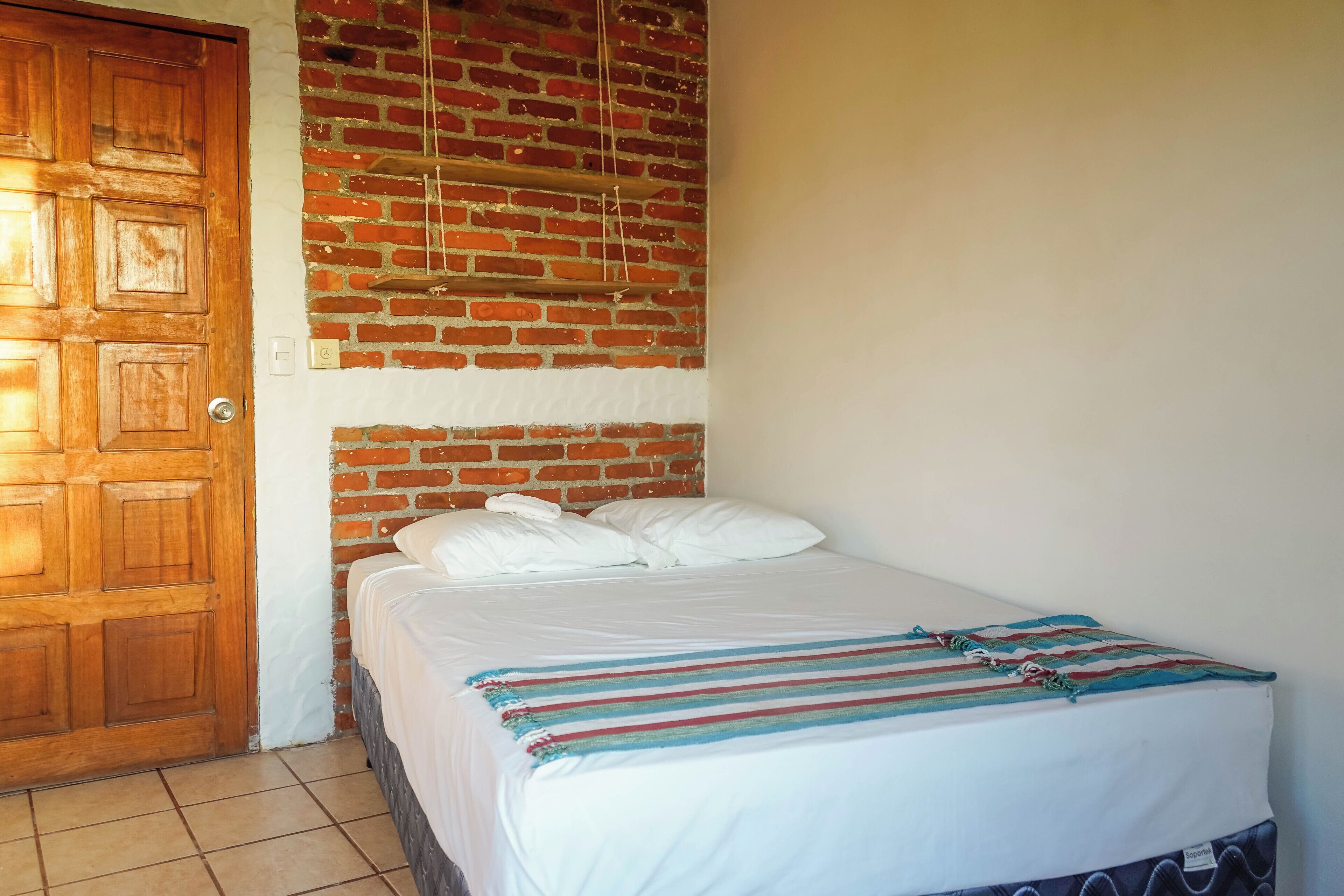 Un dormitorio con puerta de madera, pared de ladrillo, cama con ropa blanca y manta a rayas, y pared blanca.