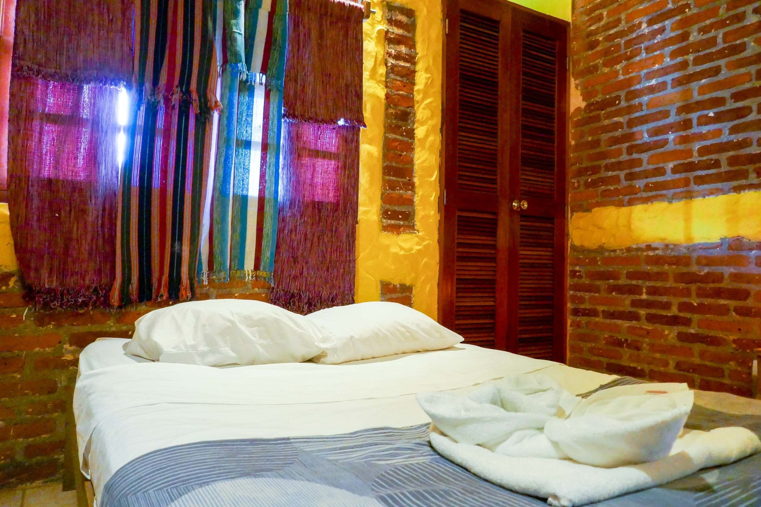 Una cama con sábanas blancas y una manta a rayas, una puerta de madera y una pared de ladrillo.
