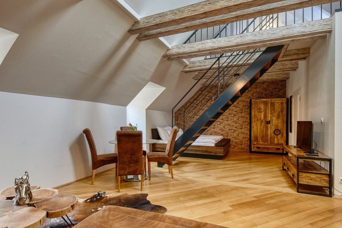 A spacious loft with exposed wooden beams, a loft bed, a staircase, and a cozy seating area with a brown sofa.