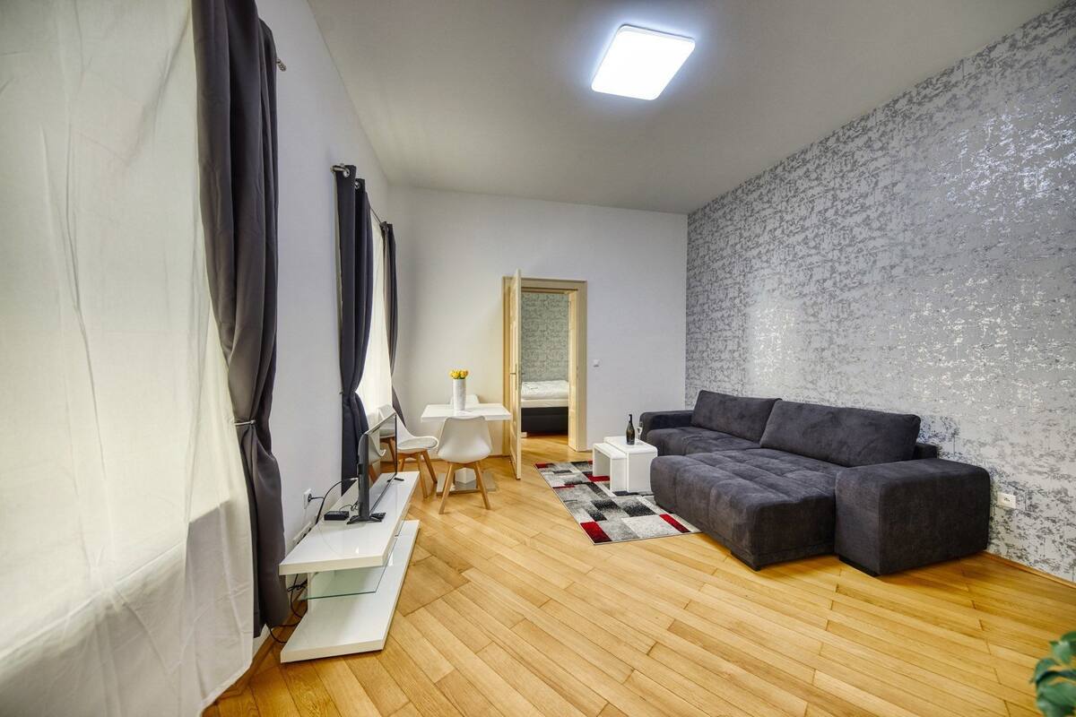 A modern living room with a grey sofa, a white side table, a glass coffee table, a white chair, and a patterned rug.