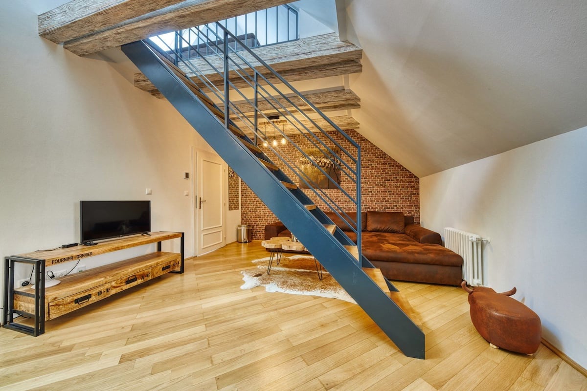 A loft-style living space with a sloped ceiling, exposed brick walls, a wooden floor, a blue staircase, a leather sofa, and a television on a wooden console.