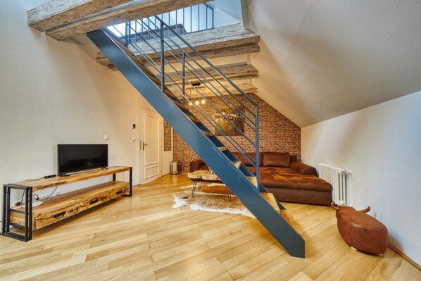 Un espacio habitable estilo loft con techo inclinado, paredes de ladrillo visto, piso de madera, una escalera azul, un sofá de cuero y un televisor sobre un mueble de madera.