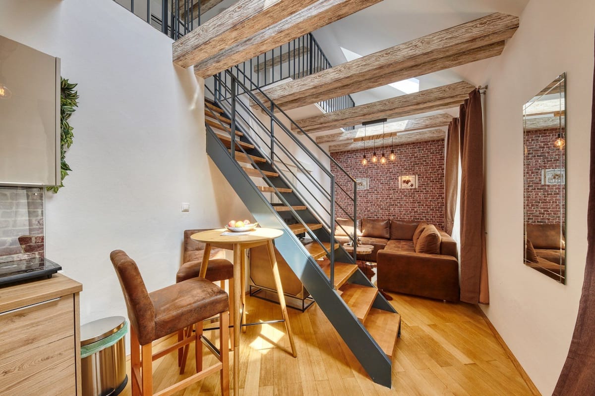 A modern loft with exposed wooden beams, a staircase, a brick wall, and a cozy seating area with a table and chairs.