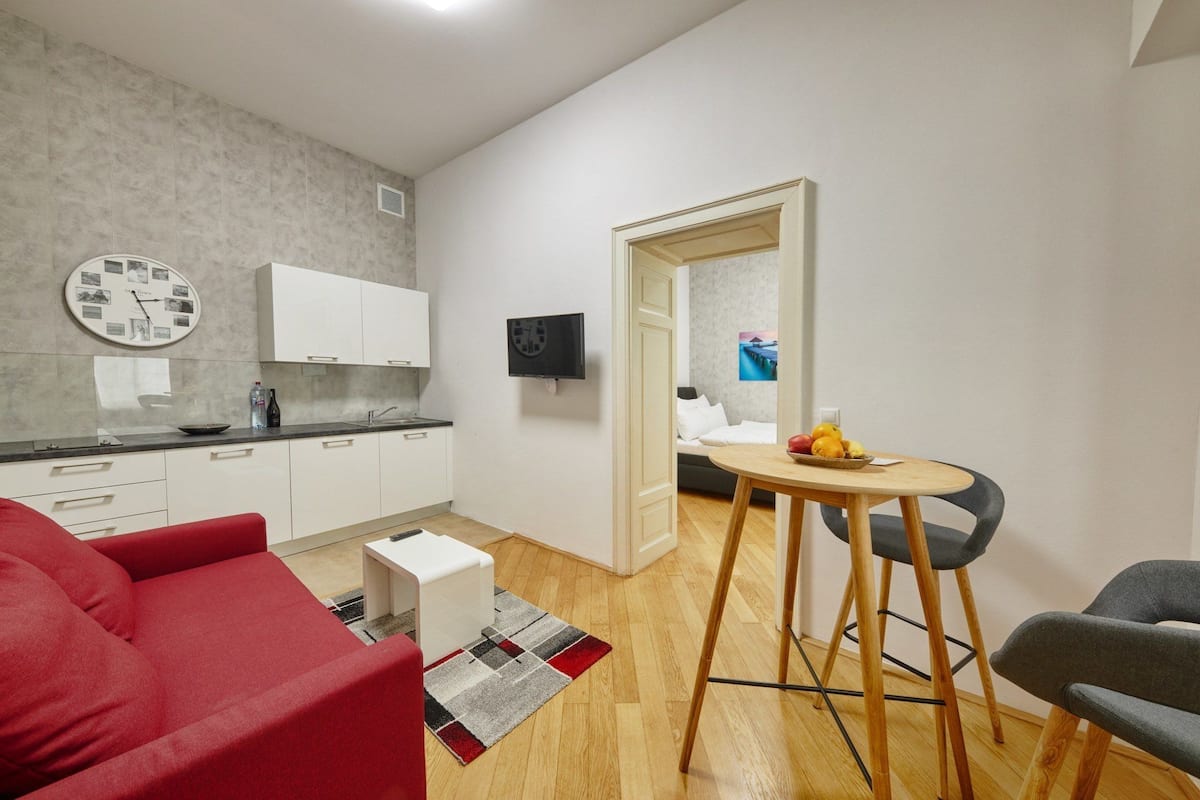 A compact living space with a kitchenette, a dining area with a small table and chairs, a red sofa, and a grey armchair.