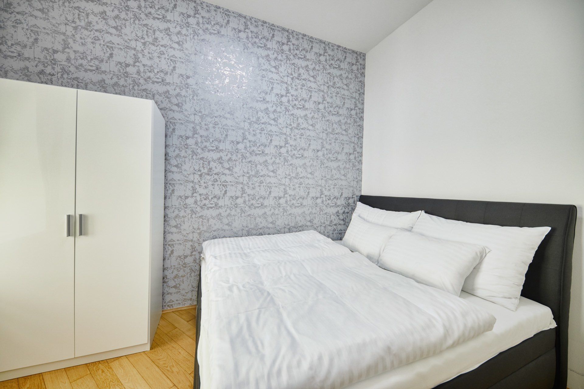 A bedroom with a bed, white bedding, a wardrobe, and a patterned wall.