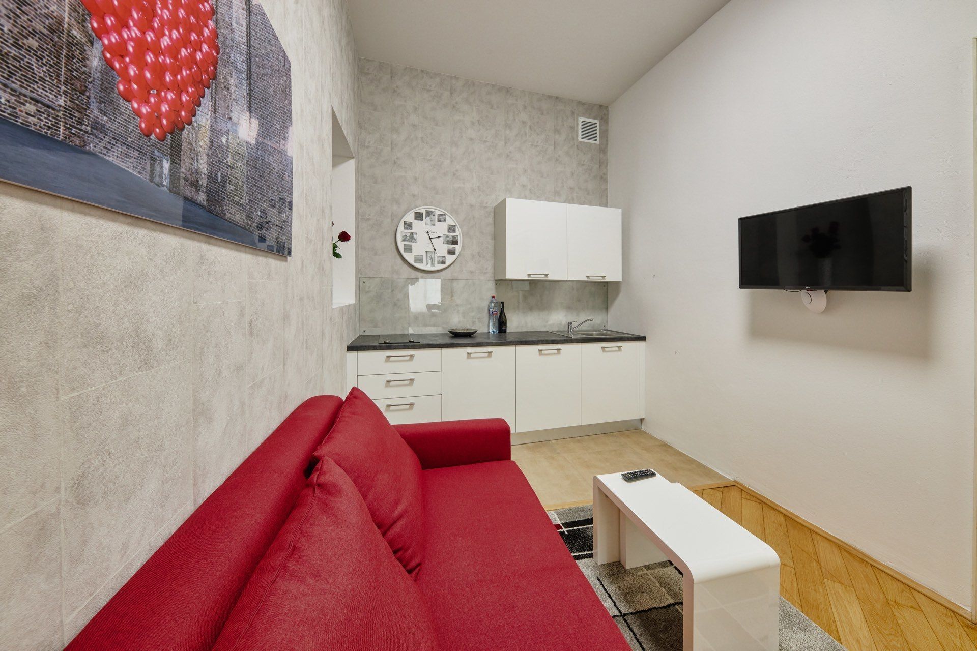 A compact living space with a kitchenette, a red sofa, a small white table, and a mounted TV.