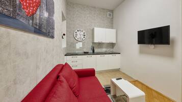 A compact living space with a kitchenette, a red sofa, a small white table, and a mounted TV.