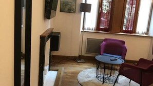 Un salon doté d’un parquet en bois, d’une table ronde, de deux chaises, d’un téléviseur fixé au mur et d’une fenêtre avec des rideaux.
