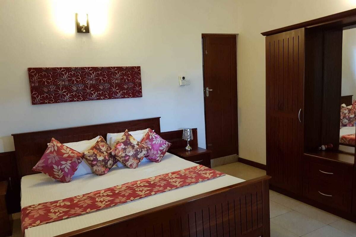 A bedroom with a wooden bed, floral-patterned pillows, a bedside table with a lamp, a wardrobe, and a wall-mounted light fixture.