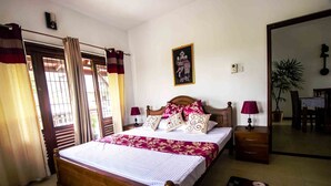 Un dormitorio con cama de madera, dos mesitas de noche, un tocador, un espejo, una puerta con cortinas y una planta en maceta.