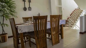 Un comedor con sillas de madera y una mesa dispuesta para cuatro personas, una escalera al fondo y una planta en maceta a la izquierda.