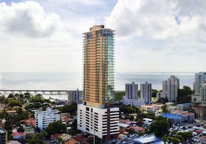 A tall building with a modern design, surrounded by other buildings and a body of water in the background.