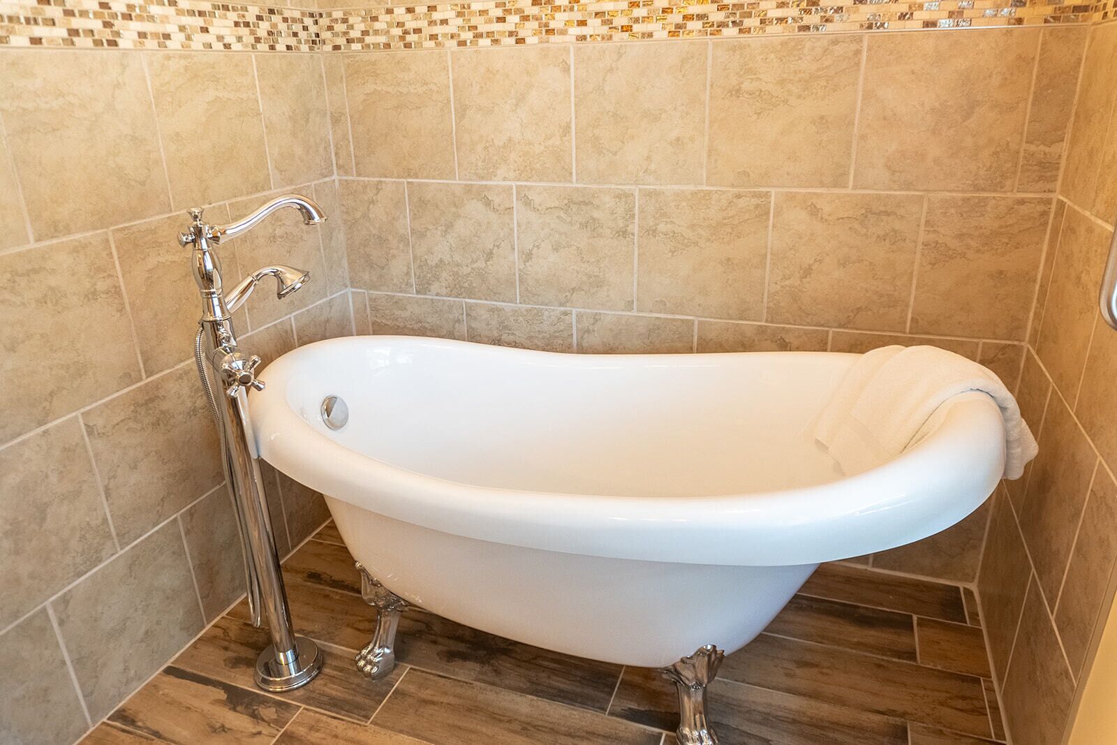 Una bañera independiente con grifo y ducha de cromo, frente a una pared de azulejos con un detalle de mosaico.