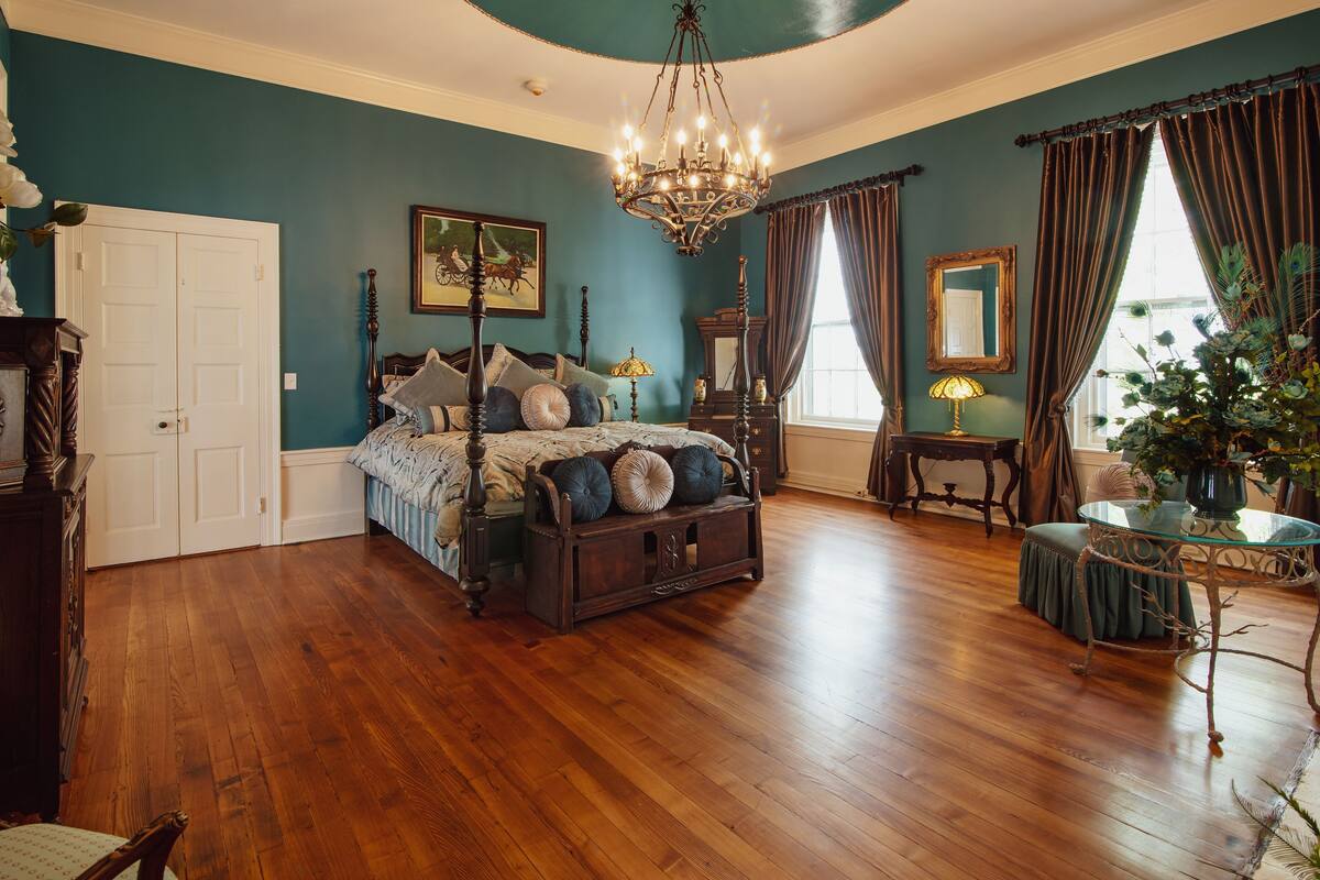 A bedroom with a wooden floor, a bed with a canopy, a chandelier, a dresser, a mirror, and a window with curtains.