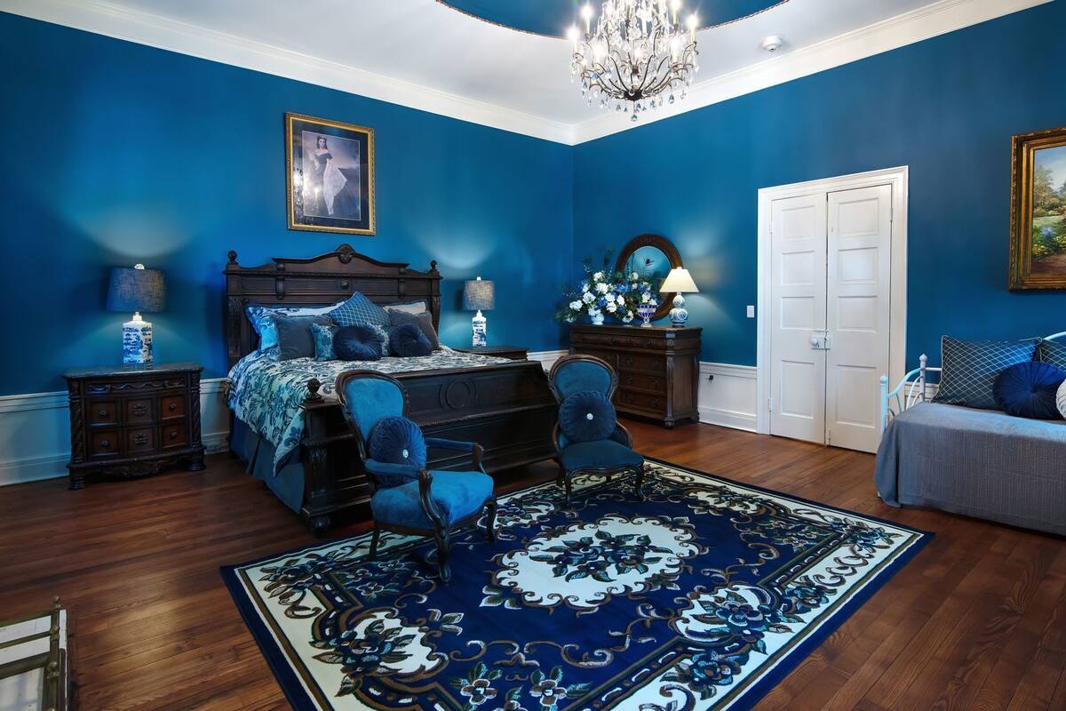 A bedroom with a large bed, a chandelier, a dresser, a chair, and a door.