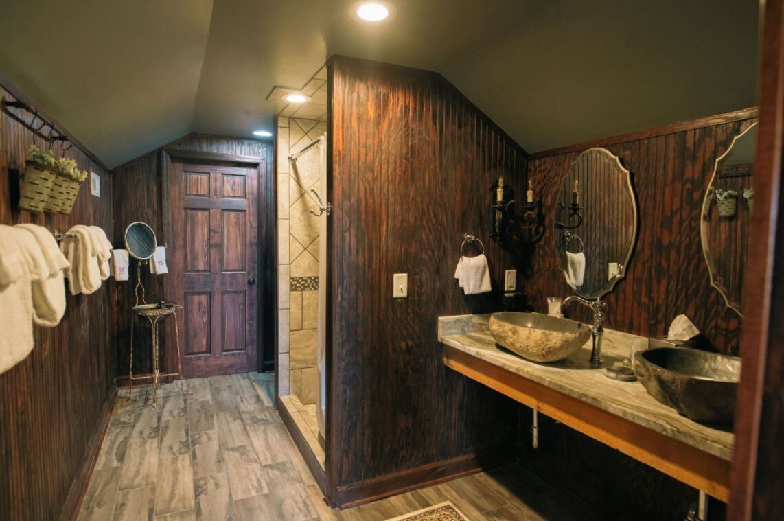 A wooden bathroom with a large mirror, two sinks, and a door leading to another room.