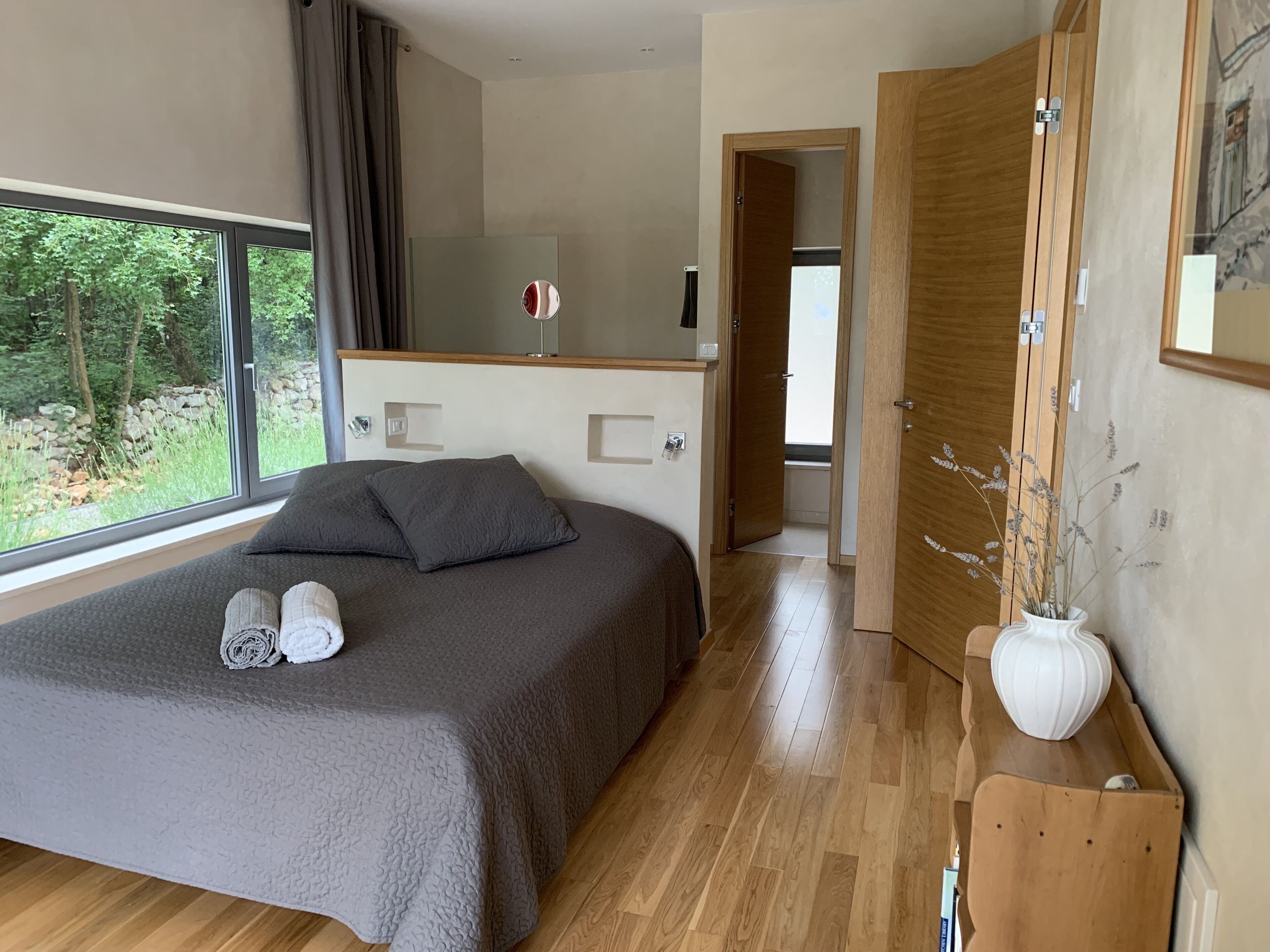 A modern bedroom with a large bed, a bedside table with a vase, and a view of greenery outside.