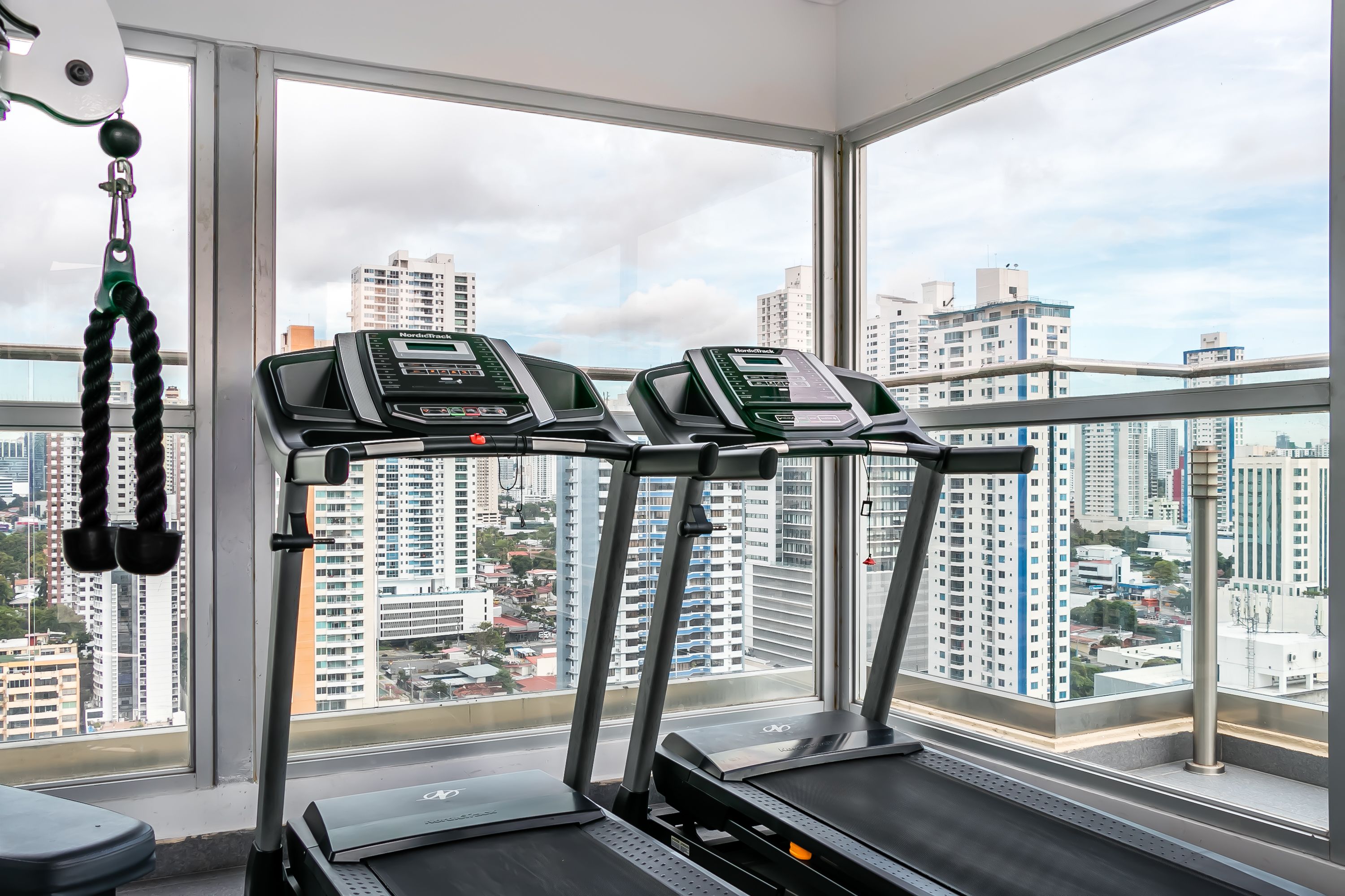 Un gimnasio con cintas de correr y máquinas de pesas, amplio ventanal con vista a la ciudad.