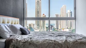 A bed with a patterned bedspread, a headboard, and pillows, with a cityscape view through a large window.
