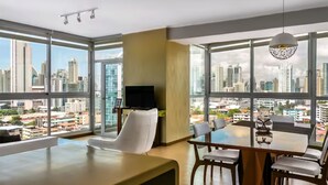 A modern hotel room with large windows offering a city view, a flat-screen TV, and a dining area with a table and chairs.