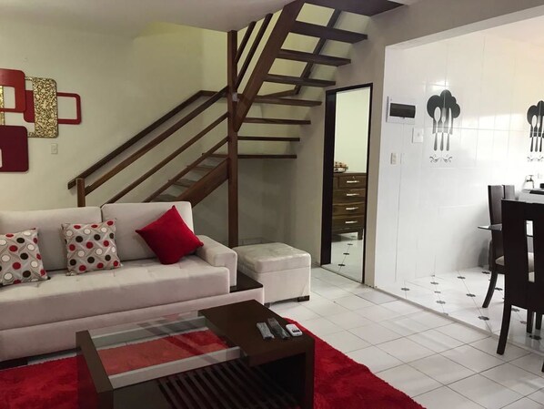 Una sala de estar moderna con una escalera, un sofá blanco con cojines estampados, un tapete rojo y una zona de comedor con una silla y una mesa.