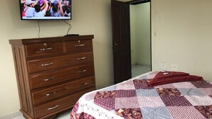 Un dormitori amb un taulell de fusta, un llit amb estorí i una televisió penjada a la paret, i una porta que dona a una altra habitació.