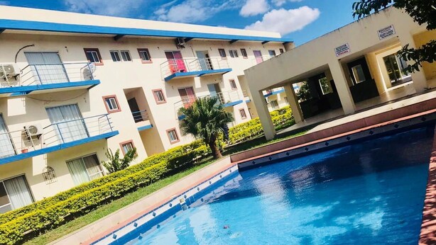 Una piscina al costat d'un edifici de diversos pisos amb balcons, un cel serè i jardineria ben cuidada.