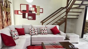 Un saló modern amb un sofà blanc, coixins vermells i amb dibuixos, una taula de centre de vidre i una escala al fons.