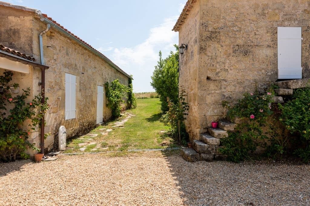 A narrow gravel path leading through a rustic stone building with white shutters, surrounded by greenery and a clear sky.