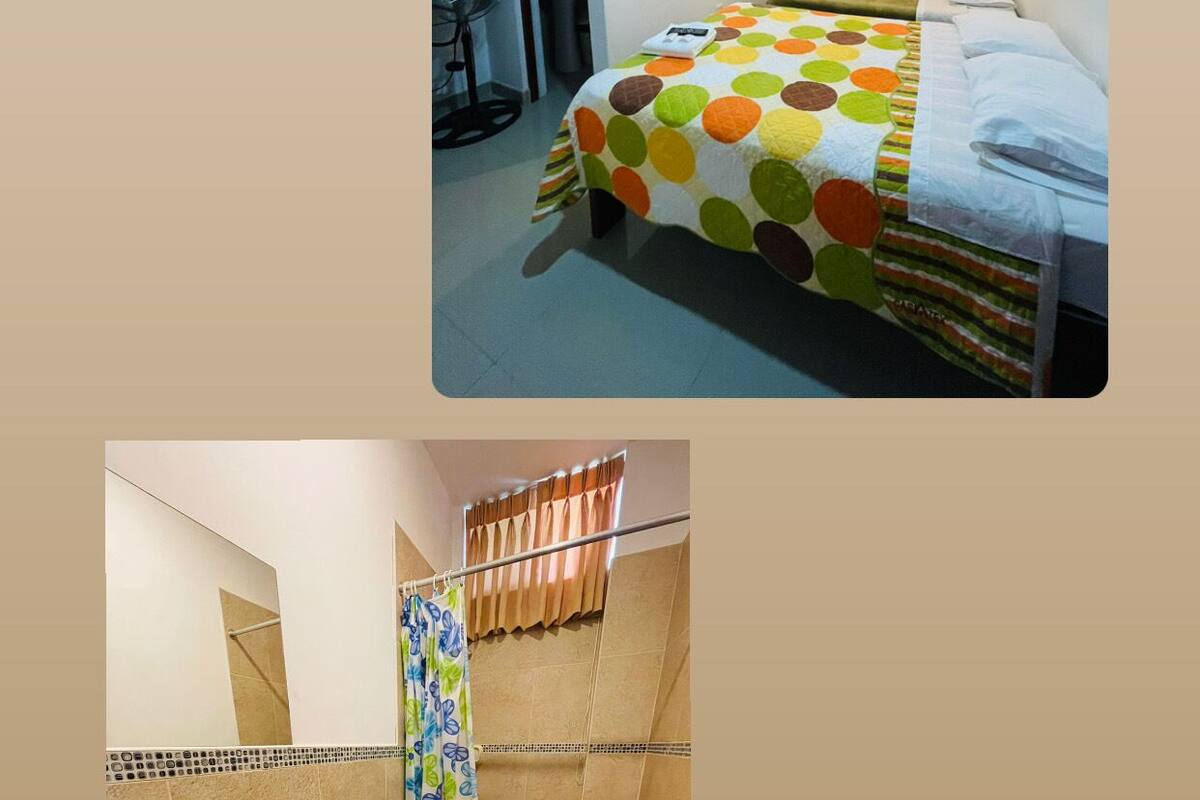 A bedroom with a bed, a bedside table, and a window with curtains. The room has a door leading to another area.