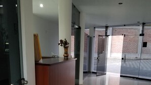Una moderna zona de recepción con mostrador de madera, un jarrón con flores y una puerta de cristal que da a un exterior de ladrillo.