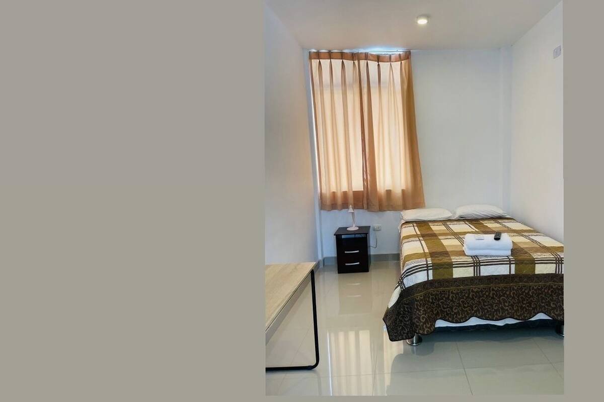 A bedroom with a bed, bedside table, and curtains. The bathroom includes a bathtub, toilet, sink, and shower curtain.